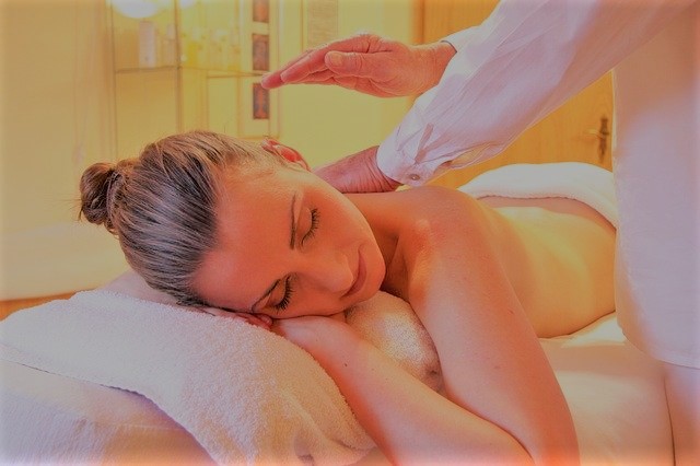 Relaxed woman lying on a massage table with eyes closed while a therapist performs an energy healing or massage session in a calm wellness setting.