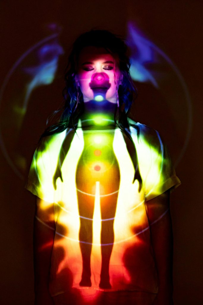 Une femme se tient dans une pièce sombre avec une projection de chakra colorée sur son corps.